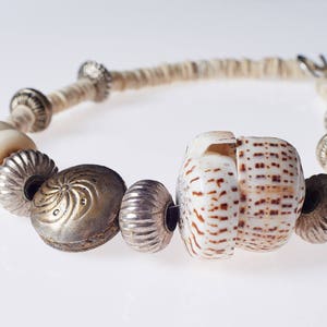 Könnte beinhalten: Eine Halskette aus weißen und braunen Muschelkernperlen mit silbernen und weißen Perlen. Die Halskette ist aus natürlichen Materialien gefertigt und hat einen rustikalen Look.