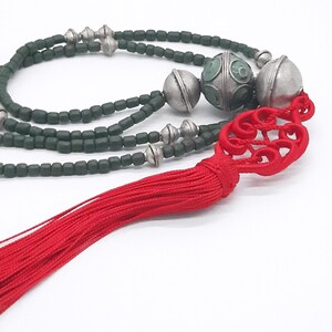 Peut inclure: Un collier composé de perles vert foncé, d'éléments argentés et de grosses perles argentées décoratives. Un pompon rouge vif avec un motif complexe ajoute une touche de couleur. Le collier est probablement fait à la main et adapté aux occasions spéciales.