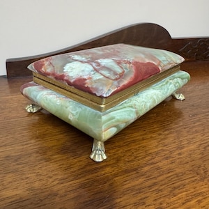 Può includere: Un portagioie rettangolare con coperchio incernierato. La scatola è realizzata in marmo verde e rosso con finiture e piedini color oro. Il marmo presenta un motivo naturale a spirale. La scatola è su una superficie di legno.