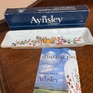 May include: Aynsley China rectangular serving dish with a floral and fruit design, gold trim, and the brand name on the bottom. The box reads "Fondé en 1775" and "Fine Anglaise Porcelain". A Somerset brochure is also present.