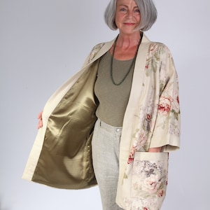 May include: A cream-coloured floral kimono with a green satin lining. The kimono has a floral print with pink, red, and green flowers. The model is wearing the kimono open, showing the lining.