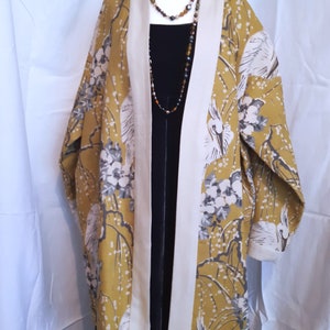 Puede incluir: Un kimono largo de estampado amarillo y blanco con cuello blanco y un vestido negro debajo. El kimono tiene un diseño floral y de pájaros.