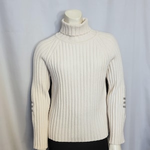 Op de afbeelding: Crèmekleurige geribbelde gebreide coltrui. De trui met lange mouwen heeft een hoge halslijn en verticale ribbels. De mouwen hebben een klein grijs ontwerp bij de elleboog. De trui wordt op een mannequin getoond.