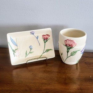 Puede incluir: Un juego de jabonera y vaso de cerámica color crema. La jabonera tiene un diseño acanalado con detalles florales. El vaso tiene un diseño acanalado a juego y detalles florales. Las flores son rosas, moradas y azules con tallos y hojas verdes.
