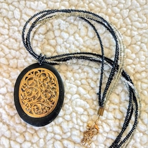 Puede incluir: Un collar de múltiples hilos con un colgante ovalado negro y un diseño de filigrana dorada. El collar tiene hilos de cuentas negras y plateadas, y un cierre dorado.