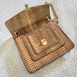 May include: A brown faux snakeskin handbag with a gold chain strap and clasp. The bag is open, revealing a smaller compartment inside. The bag is square-shaped and has a textured pattern.