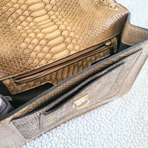 May include: Open view of a tan faux snakeskin handbag. The bag features a gold chain strap, a gold clasp, and multiple interior compartments, including a zippered pocket. The interior lining is a darker shade of brown.