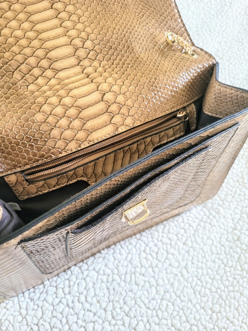 May include: Open view of a tan faux snakeskin handbag. The bag features a gold chain strap, a gold clasp, and multiple interior compartments, including a zippered pocket. The interior lining is a darker shade of brown.