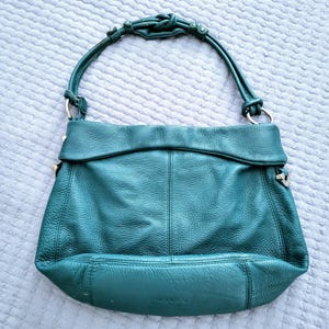May include: A teal leather handbag with a knotted handle and silver-toned hardware. The bag has a fold-over flap and a textured surface. The bag is a medium size and has a rounded bottom.