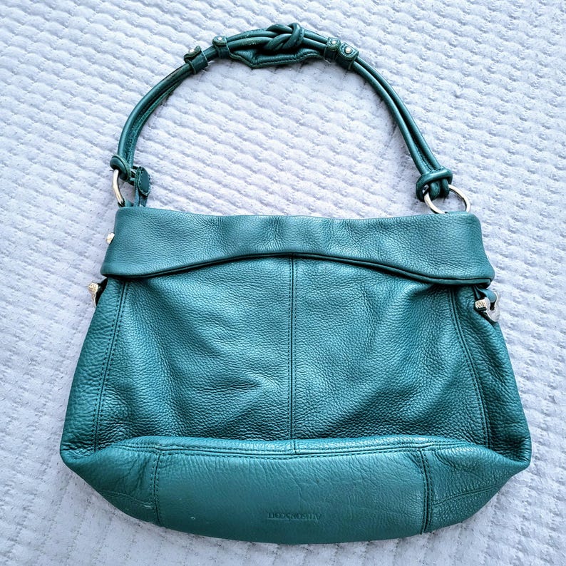May include: A teal leather handbag with a knotted handle and silver-toned hardware. The bag has a fold-over flap and a textured surface. The bag is a medium size and has a rounded bottom.