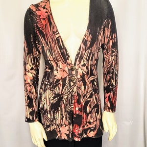 May include: A long-sleeved cardigan with a V-neck and a floral pattern in shades of black, peach, and cream. The cardigan has two decorative buttons and is displayed on a mannequin. The bottom half of the mannequin is wearing black pants.