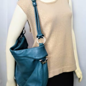 May include: Teal leather shoulder bag with a rounded shape and a fold-over flap. The bag features a silver-tone ring and hardware on the strap and body. The strap is adjustable. The bag is shown on a mannequin wearing a gold-colored top and black skirt.