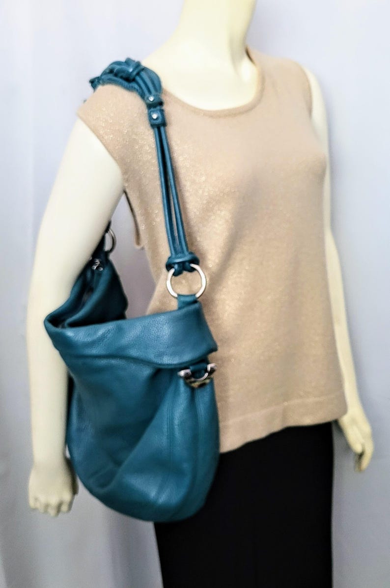 May include: Teal leather shoulder bag with a rounded shape and a fold-over flap. The bag features a silver-tone ring and hardware on the strap and body. The strap is adjustable. The bag is shown on a mannequin wearing a gold-colored top and black skirt.