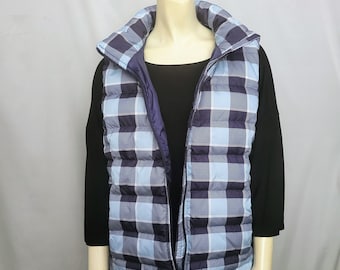 May include: A reversible puffer vest with a blue and navy plaid pattern. The vest has a high collar and a full-length zipper. The vest is displayed on a mannequin, over a black long-sleeve shirt and black pants.