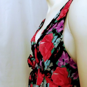 Könnte beinhalten: Ein ärmelloses Kleid mit V-Ausschnitt und einem lebendigen Blumenmuster. Das Kleid zeigt große rote, lila und blaue Blumen vor einem schwarzen Hintergrund. Der Stoff wirkt leicht und fließend, mit einem gerafften Detail an der Taille.