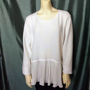 Puede incluir: Una blusa blanca de manga larga con cuello redondo y detalle plisado en el dobladillo. La prenda se exhibe en un maniquí sobre un fondo verde. Las mangas son largas y la tela parece ser un material suave.