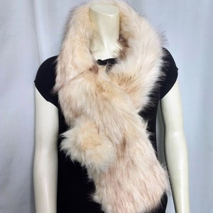 Puede incluir: Bufanda de piel sintética en tonos crema y marrón claro, colocada alrededor del cuello de un maniquí. La bufanda tiene una textura esponjosa y un diseño en capas, creando un aspecto lujoso. El fondo es una cortina blanca lisa.