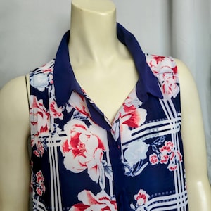 May include: Sleeveless blouse with a navy blue collar and floral print. The blouse features a navy blue background with a pattern of white and red flowers. The collar is a solid navy blue color. The blouse is buttoned down the front.