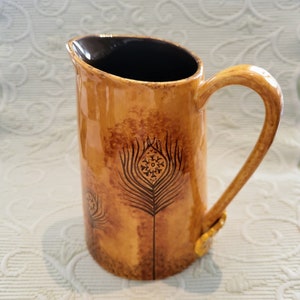 May include: A ceramic pitcher with a brown exterior and a dark brown interior. The pitcher features a decorative peacock feather design in black. The handle and base have a gold-colored accent.