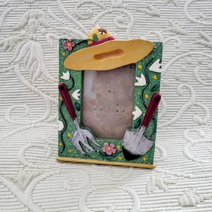 May include: A decorative photo frame with a gardening theme. The frame is green with a yellow hat at the top and gardening tools on the sides. The frame has a rectangular opening for a photo and is decorated with flowers.
