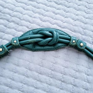 May include: Teal leather handbag strap with a knotted center detail. The strap features two parallel cords on each side, secured with small metal rivets. The background is a light blue quilted fabric.