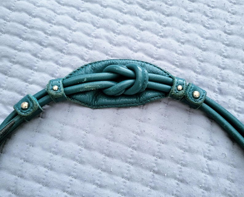 May include: Teal leather handbag strap with a knotted center detail. The strap features two parallel cords on each side, secured with small metal rivets. The background is a light blue quilted fabric.