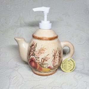 May include: A cream-colored ceramic soap dispenser shaped like a teapot with a white plastic pump. The body features a painted design of wheat stalks and fruit in shades of brown, orange, and yellow. A round tag reads Crazy Mountain Very Special Gifts.