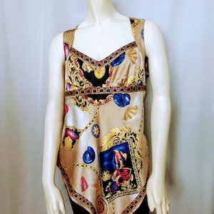 以下が含まれることがあります： ゴールドのチェーン、青い貝殻、人物の額入り画像が特徴の装飾プリントが施されたベージュのタンクトップ。 トップスはスクエアネックラインとワイドストラップです。 プリントは赤と黒のボーダーで縁取られています。