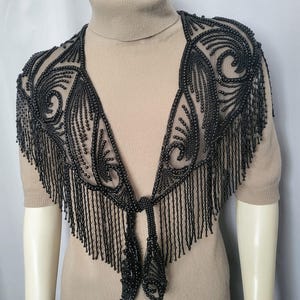 May include: A black beaded shawl featuring intricate swirling patterns and long fringe detailing. The shawl is constructed from sheer mesh fabric adorned with dense black beads, forming a V-shaped neckline that ties at the center front with beaded ends.