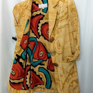 May include: Tan blazer with a patterned exterior and a vibrant interior lining. The lining features a bold design with red, teal, and black shapes. The blazer has decorative buttons and a pocket.