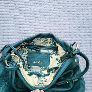 May include: Teal leather handbag with silver hardware. The interior features a floral pattern lining and a zippered pocket. The brand name "Allison Scott" is visible on a leather tag.