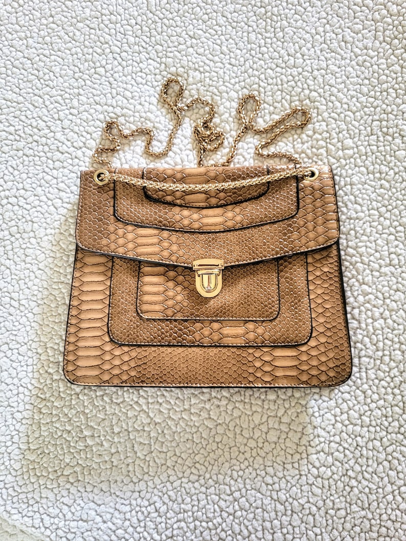 May include: A tan handbag with a snakeskin pattern and a gold chain strap. The bag has a rectangular shape with a flap closure and a gold clasp. The bag is sitting on a white, textured surface.
