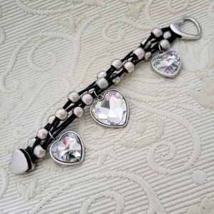 Puede incluir: Una pulsera de múltiples hilos con cordones negros, cuentas plateadas y tres dijes en forma de corazón. Los dijes están incrustados con cristales transparentes. La pulsera tiene un cierre plateado en forma de corazón.