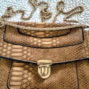 May include: A brown snakeskin-patterned handbag with a gold chain strap and a gold clasp. The bag has a flap closure and a small pocket on the front. The bag is a neutral color and could be used as a shoulder bag or crossbody bag.