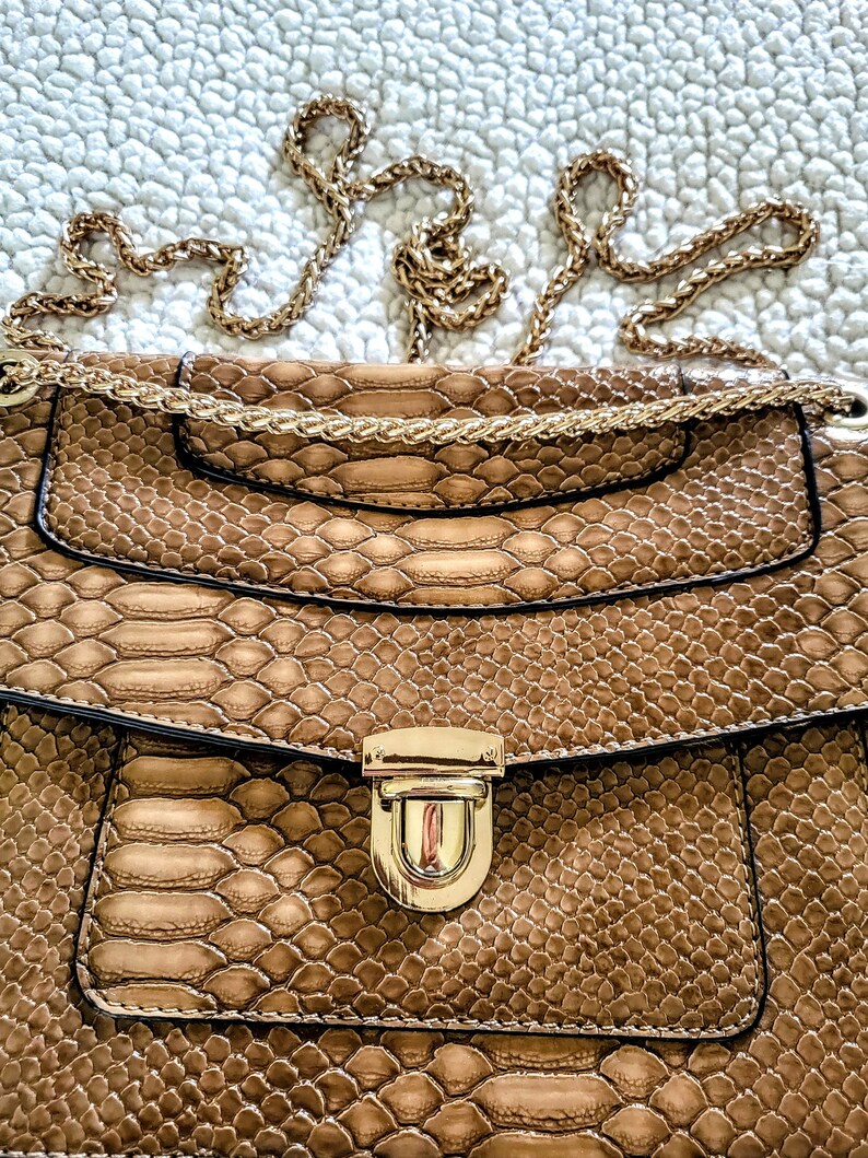 May include: A brown snakeskin-patterned handbag with a gold chain strap and a gold clasp. The bag has a flap closure and a small pocket on the front. The bag is a neutral color and could be used as a shoulder bag or crossbody bag.