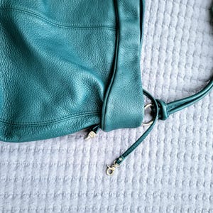 May include: Teal leather shoulder bag with a textured finish. The bag features a long strap, metal hardware, and a clasp. The bag is shown on a light gray textured surface.