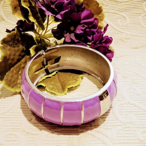 Puede incluir: Una pulsera ancha y articulada con un acabado de esmalte lavanda y detalles metálicos plateados. La pulsera tiene un diseño segmentado y se muestra con flores moradas y hojas verdes.