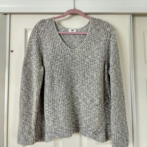 Pode incluir: Um suéter de malha cinza e branco salpicado com decote em V. O suéter está pendurado em um cabide rosa contra uma porta branca. A etiqueta da marca Old Navy é visível na gola. O suéter tem mangas compridas.