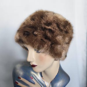 May include: A brown fur hat with a rounded crown. The hat is made of soft, textured fur in shades of brown and tan. The hat is displayed on a mannequin with a blue and silver painted bust.