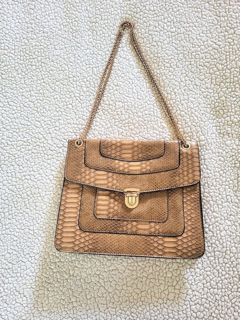 May include: A tan faux snakeskin handbag with a gold chain strap. The rectangular bag has a flap closure with a gold-tone clasp. The bag's design features layered panels and a textured pattern.