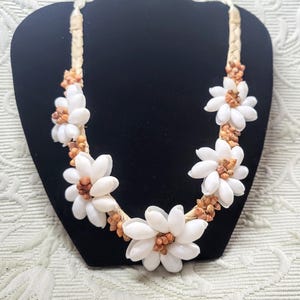 Op de afbeelding: Een handgemaakte ketting met witte schelpen bloemhangers en kleine bruine accenten. De ketting is geregen op een geweven beige koord en wordt gepresenteerd op een zwarte sieradenstandaard. Een uniek, op het strand geïnspireerd accessoire.
