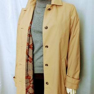 May include: A tan trench coat with a collared neckline and button closures. The coat has a patterned lining and is worn over a gray turtleneck sweater. The coat's sleeves have buttoned cuffs.