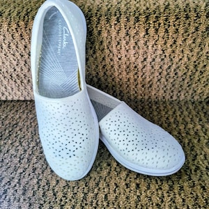 May include: A pair of white slip-on shoes with a floral pattern of cut-out designs. The shoes have a white sole and the brand name "Clarks Cloudsteppers" is visible on the insole. The shoes are on a patterned brown surface.