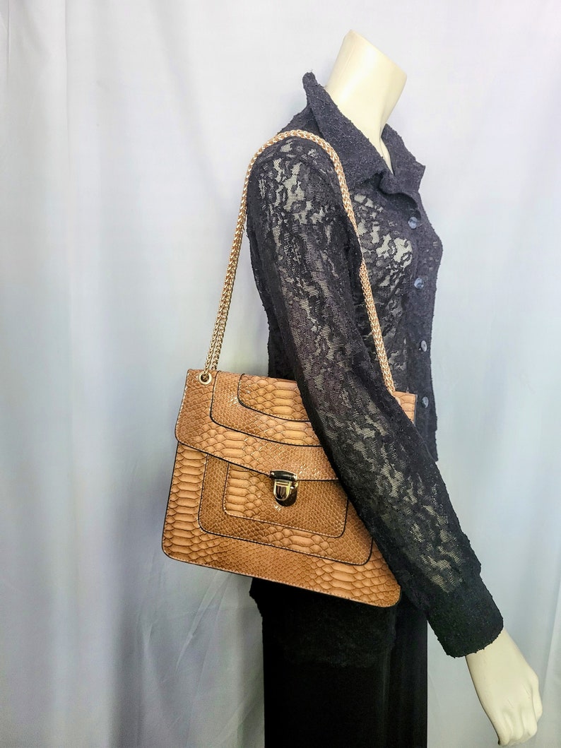 May include: A tan handbag with a snakeskin pattern and a gold chain strap. The bag has a rectangular shape with a flap closure and a gold-colored clasp. The bag is carried over the shoulder.