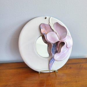 Peut inclure: Un miroir mural rond en céramique blanche avec un motif floral sculpté en trois dimensions dans des tons de rose, violet et bleu. Le miroir est situé au centre du cercle en céramique blanche.