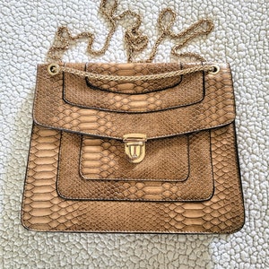 May include: A tan handbag with a snakeskin pattern and a gold chain strap. The bag has a rectangular shape with a flap closure and a gold-colored clasp. The bag is sitting on a white, textured surface.