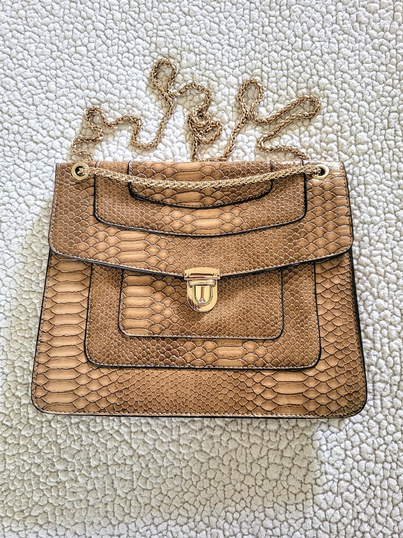May include: A tan handbag with a snakeskin pattern and a gold chain strap. The bag has a rectangular shape with a flap closure and a gold-colored clasp. The bag is sitting on a white, textured surface.