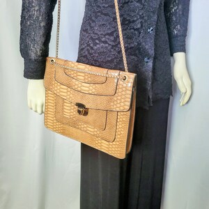 May include: A tan faux snakeskin shoulder bag with a gold chain strap. The bag has a rectangular shape with a flap closure and a decorative lock. The bag is held by a mannequin wearing a black lace top and black pants.
