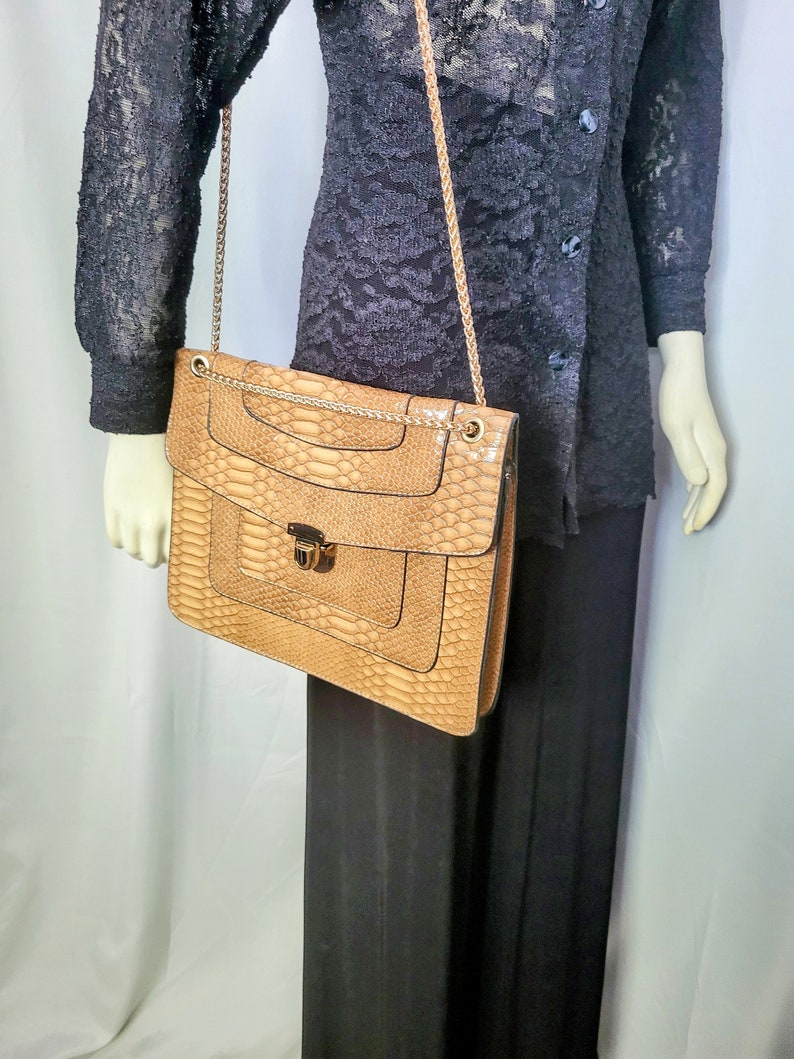 May include: A tan faux snakeskin shoulder bag with a gold chain strap. The bag has a rectangular shape with a flap closure and a decorative lock. The bag is held by a mannequin wearing a black lace top and black pants.