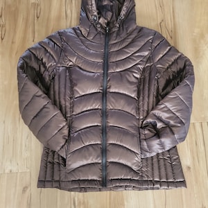 Puede incluir: Una chaqueta acolchada marrón con capucha y cremallera negra completa. La chaqueta tiene un acabado brillante y metálico y está diseñada con patrones de costura horizontales y verticales. Las mangas son largas y la chaqueta parece estar hecha de un material ligero.